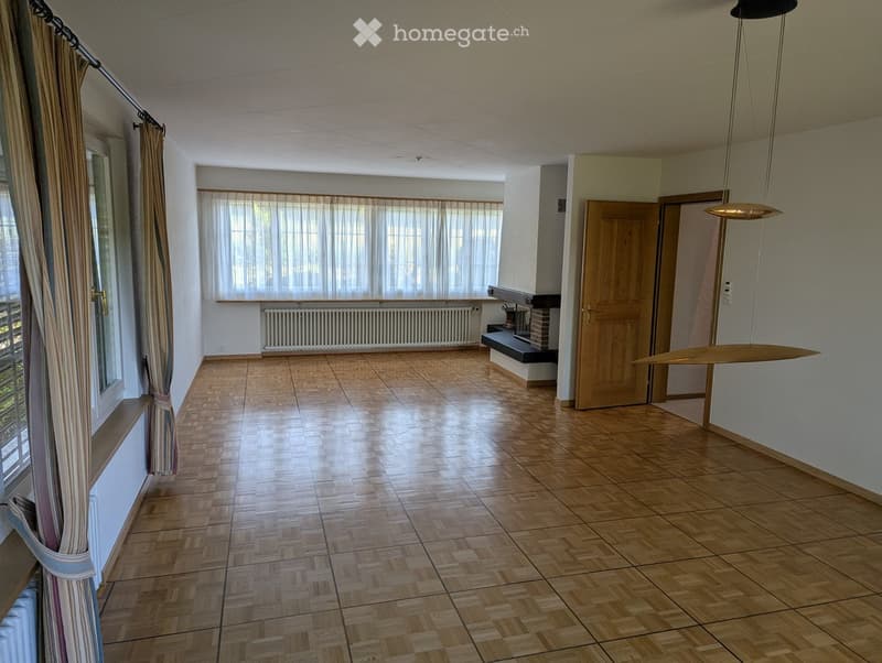 Empty room with wooden flooring, large windows with curtains, radiator, and a hanging light fixture.