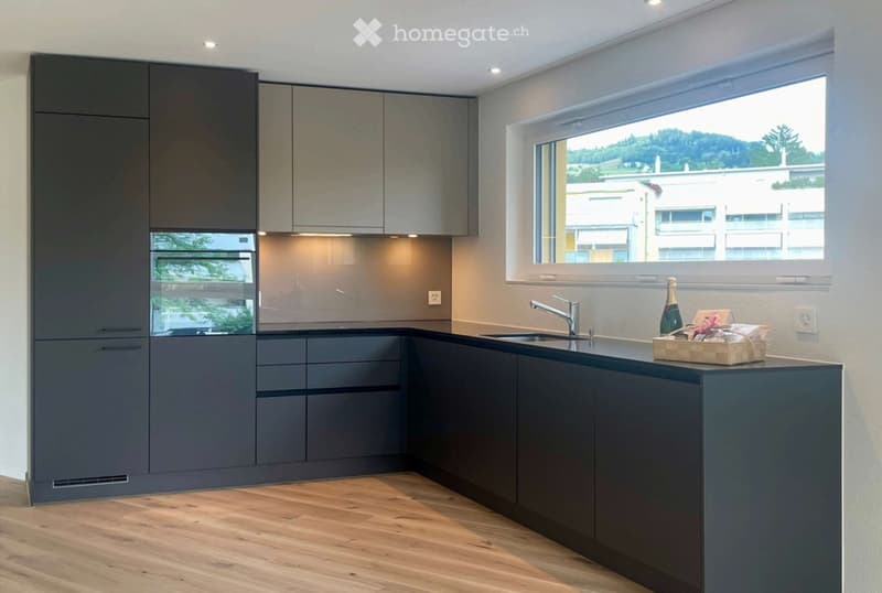 Modern kitchen with dark grey cabinets, wooden flooring, and a large window overlooking a scenic view.