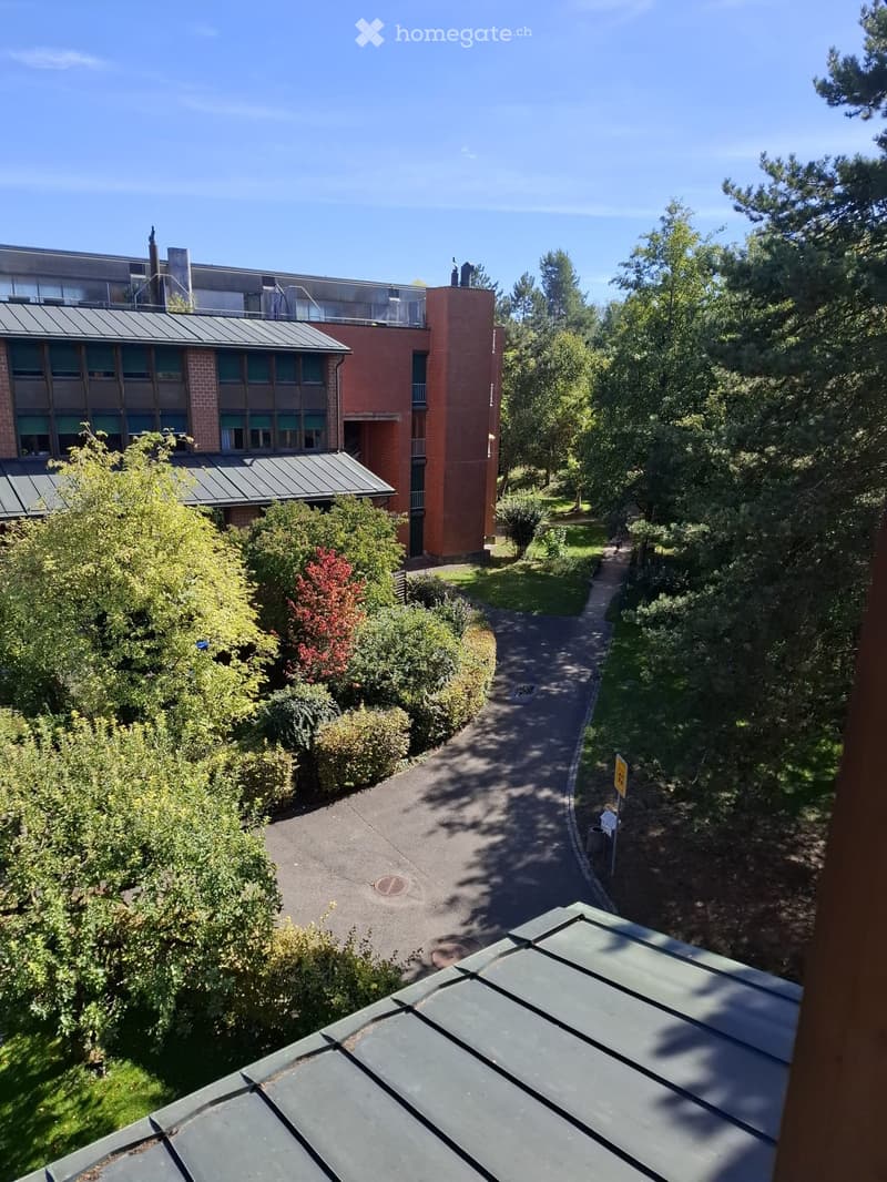 A modern multi-story building with red brick and grey metal roofing, surrounded by greenery and pathways.