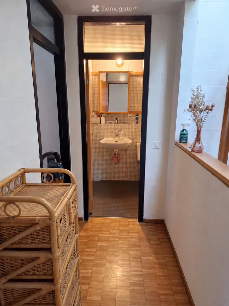 A narrow hallway with wooden flooring, white brick walls, and a small bathroom visible at the end. The hallway features a wicker basket on the left and a vase with dried flowers on the windowsill to the right.