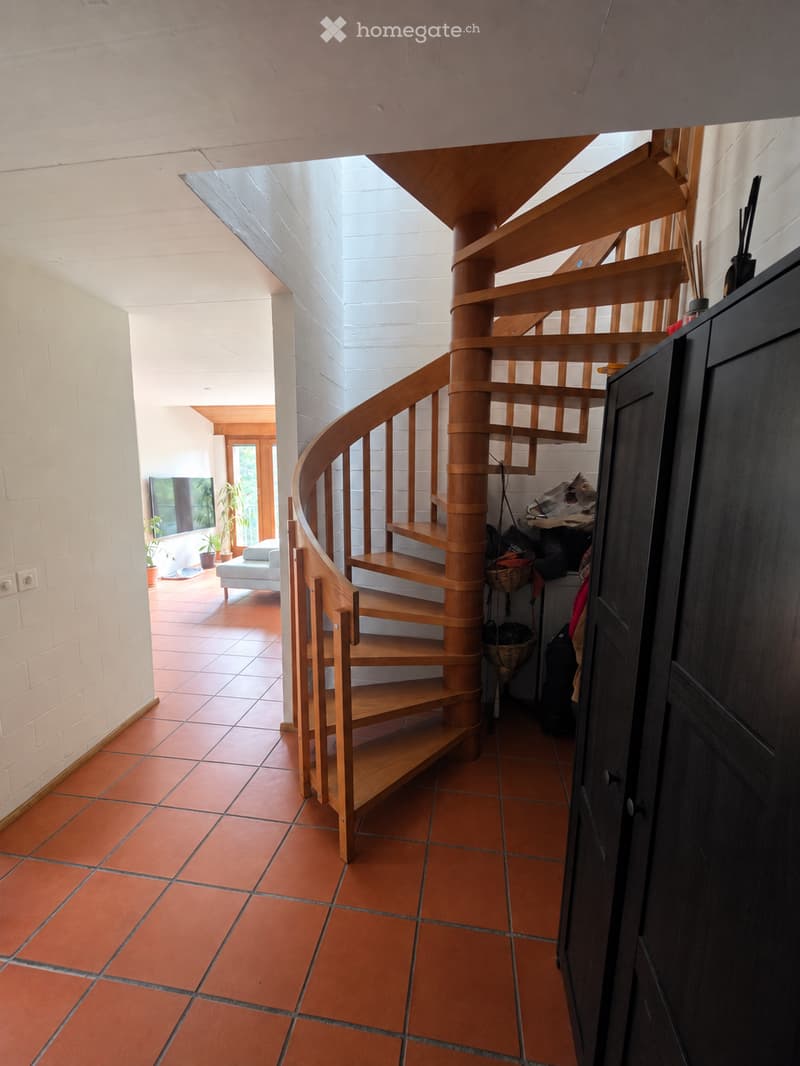 Spiral wooden staircase with open wooden railings, terracotta tiled floor, white brick walls, and a view into a living area with a sofa, TV, and potted plants.