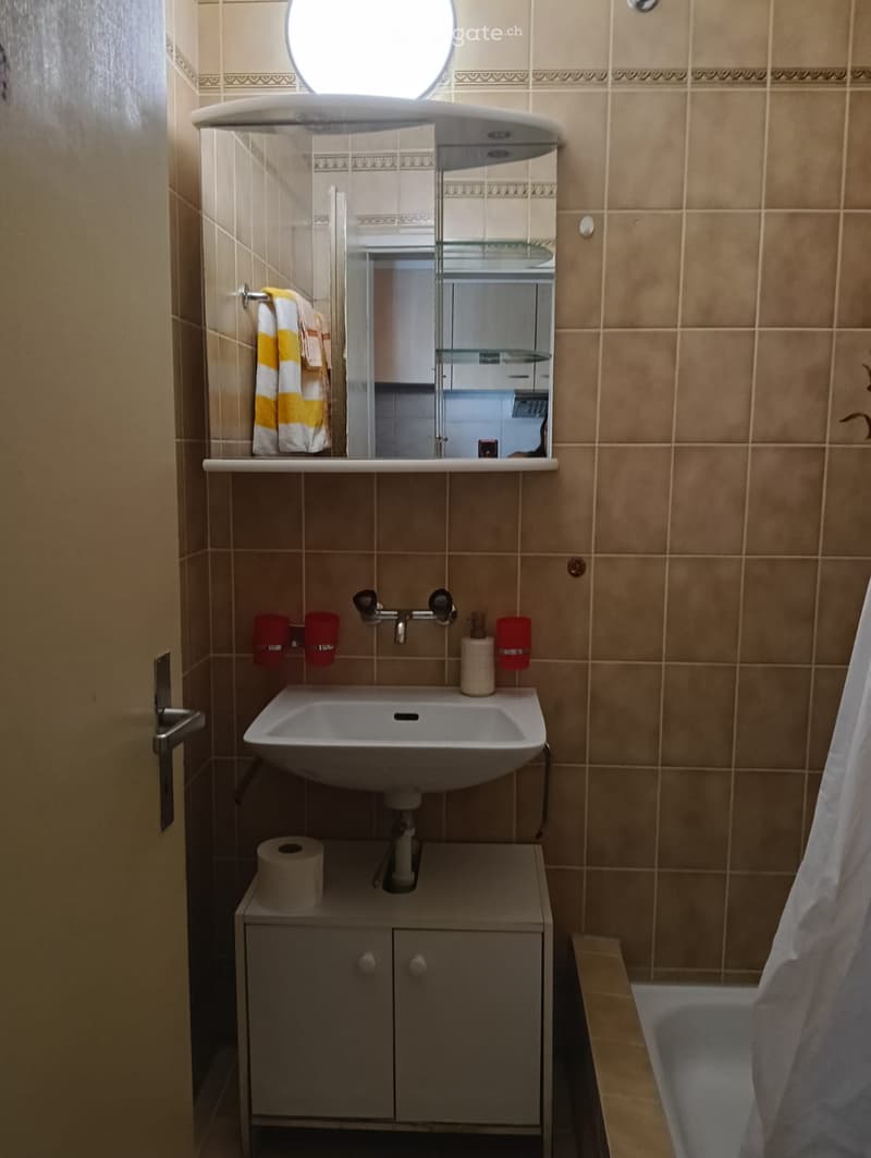 Bathroom with beige tiled walls, a white sink, a mirror with a glass shelf, a small cabinet, and a bathtub with a curtain.