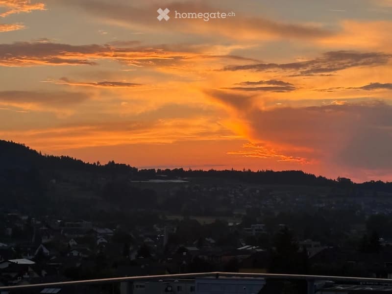 View from a balcony or terrace showing a cityscape at sunset with a hill in the background.