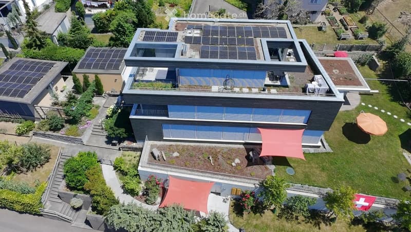 A modern two-story house with solar panels on the roof, surrounded by a well-maintained garden with patio areas and outdoor furniture.