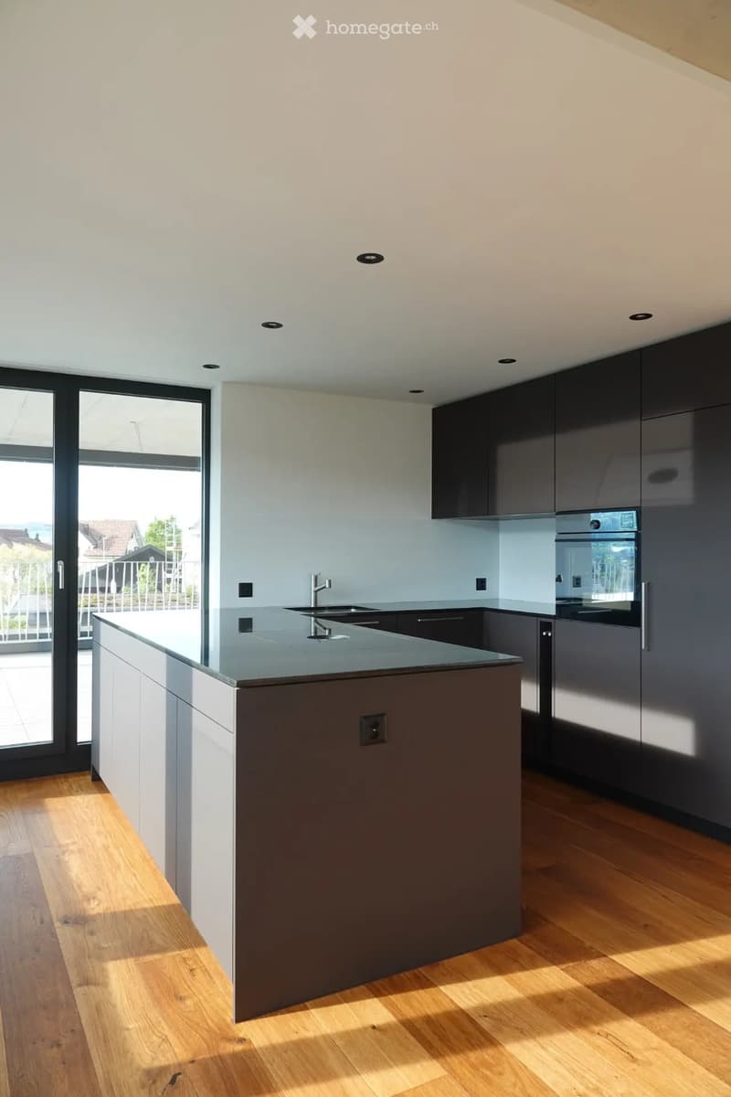 Modern kitchen with a central island, sleek grey cabinets, and a stainless steel sink and oven. The space features wooden flooring and large windows offering a view of the outdoor area.