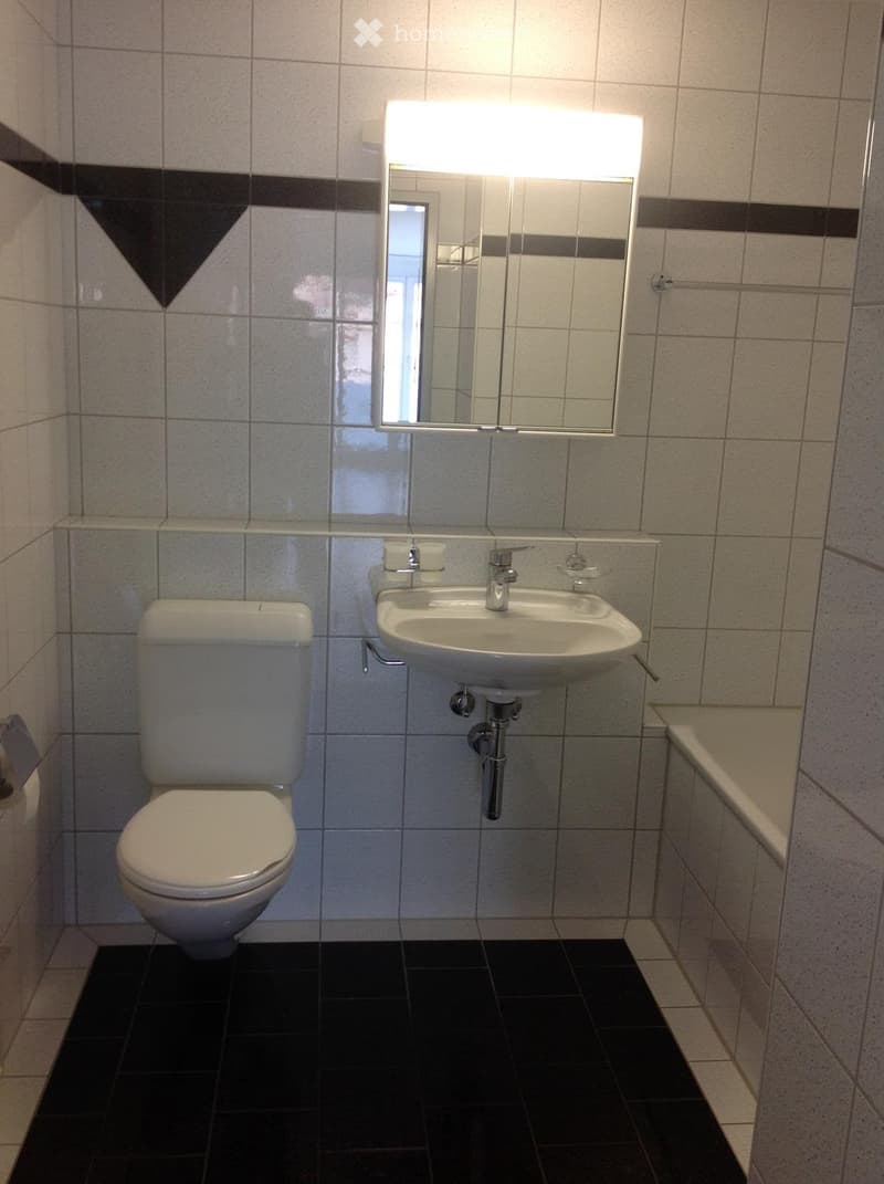 Bathroom with a toilet, sink, and mirror. The walls are tiled with a combination of light and dark tiles, and the floor is black tiled.