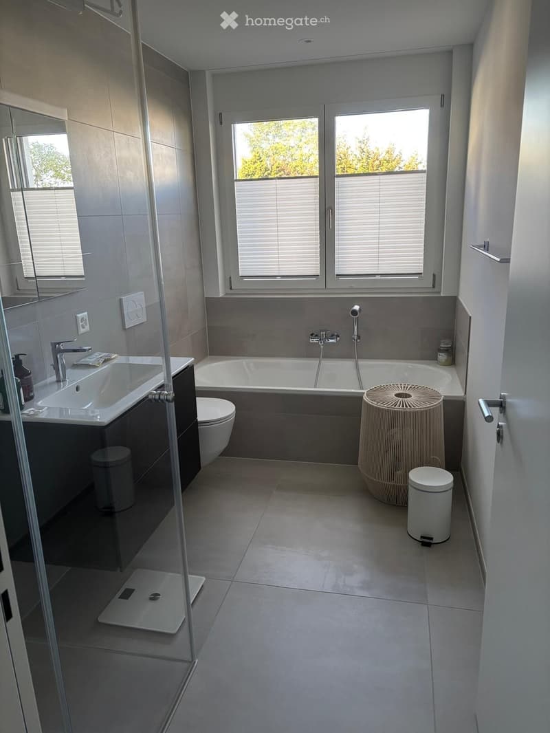 Modern bathroom with a bathtub, sink, toilet, and a wicker stool near the window. Features large windows with blinds, a glass shower stall, and a sleek design.