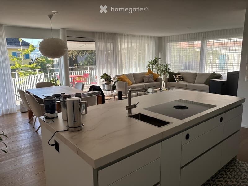 Open-plan living and dining area with a modern kitchen island, sofa, dining table, and large windows leading to a balcony.