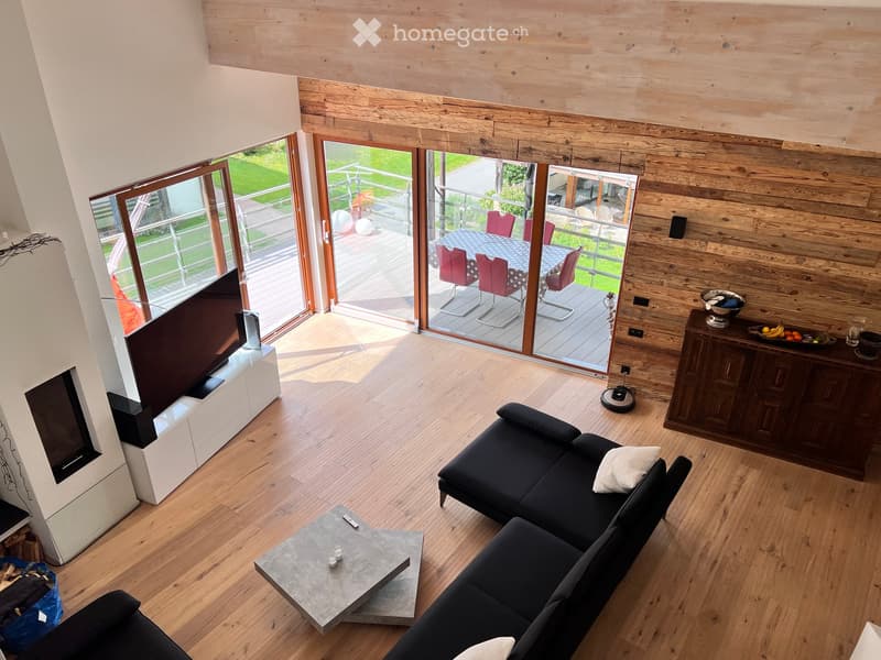 Modern living room with wooden flooring, large windows, and a balcony. Features a black sectional sofa, a grey coffee table, a TV on a white stand, and a wooden cabinet with a fruit bowl.