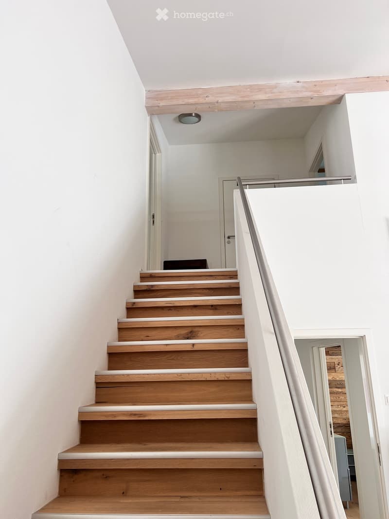 Wooden staircase with white trim, modern railing, and recessed ceiling light. The walls are white, and the ceiling has a sloped design.