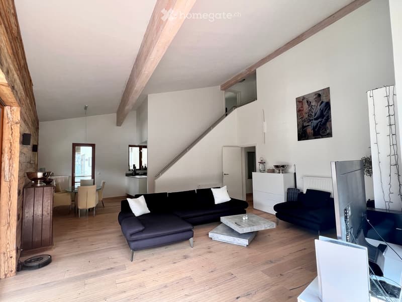 Modern living room with exposed wooden beams, hardwood flooring, a sectional sofa, coffee table, dining area with chairs and table, and a TV screen.