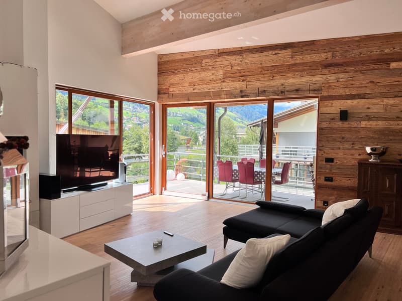 Modern living room with large windows offering a scenic mountain view, a black sectional sofa, a coffee table, a TV on a white stand, and a balcony with red chairs and a table.