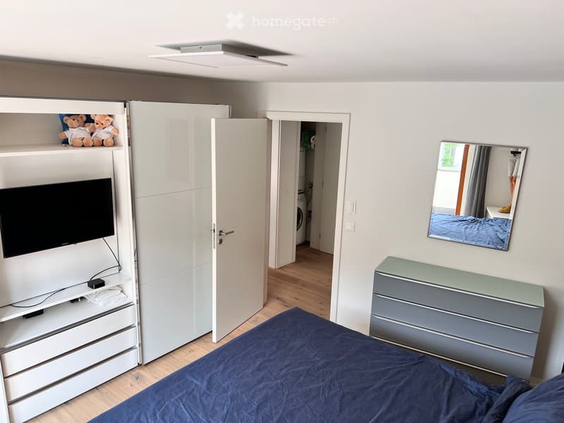Modern bedroom with a blue bedspread, white wardrobe, TV on a white stand, mirror, and nightstand with drawers.