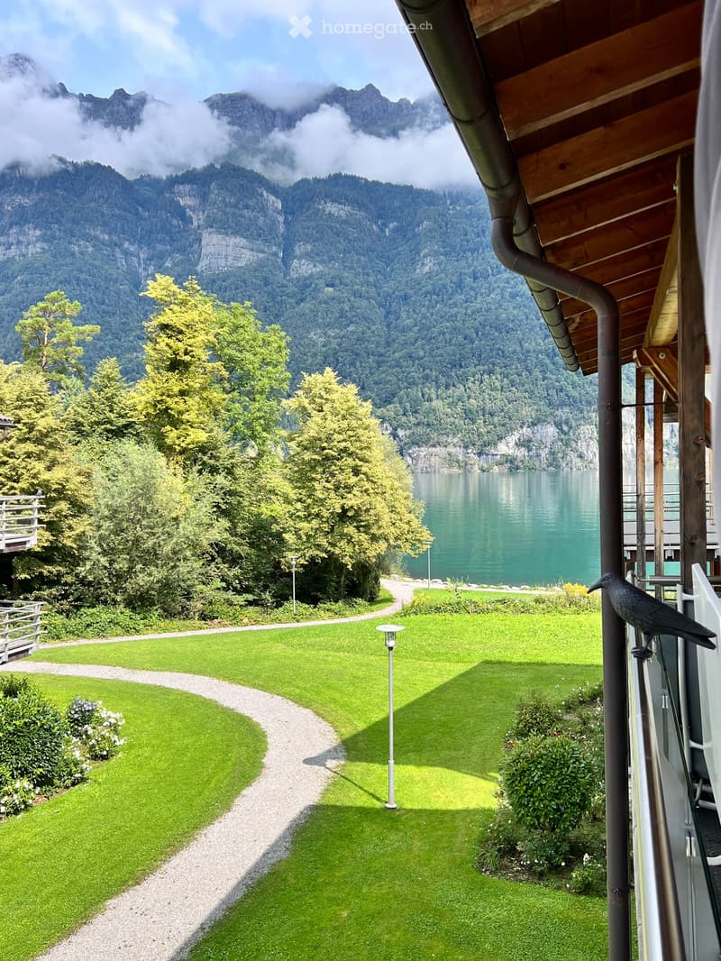 Lush green lawn with a curved gravel path, surrounded by trees and bushes, overlooking a serene lake and mountain backdrop.