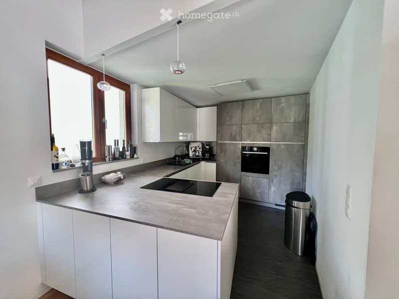 Modern kitchen with white cabinets, grey countertops, built-in oven, induction cooktop, and various appliances. Features a large window with wooden frame and pendant lighting.