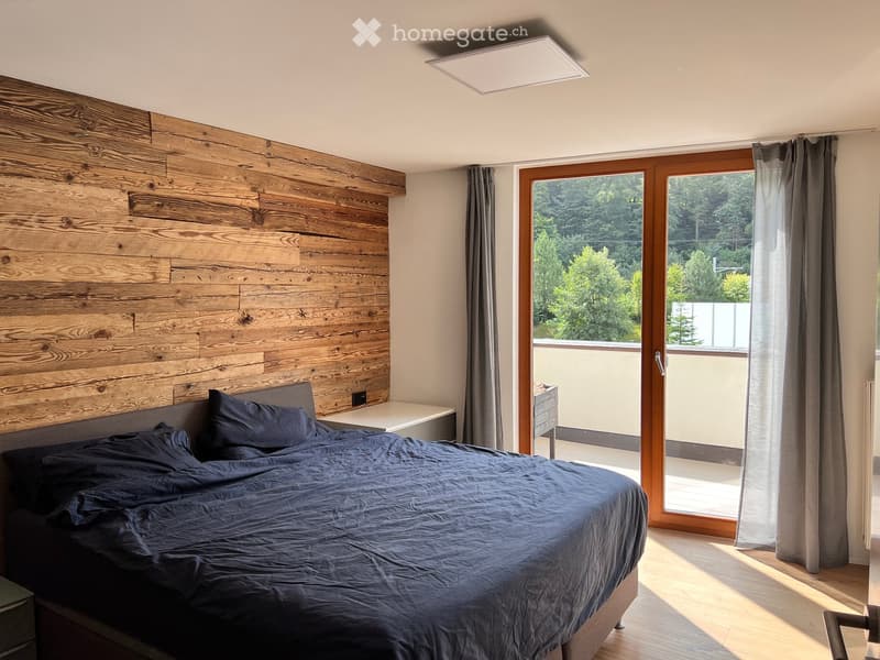Modern bedroom with a wooden accent wall, large window with view, bed with dark bedding, nightstand, and curtains.