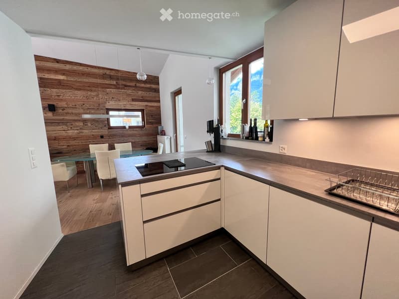 Modern kitchen with white cabinets, wooden accents, and a glass dining table with chairs. Features a dishwasher, countertop, and hanging light fixtures.