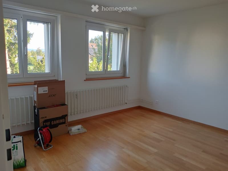 Empty room with wooden flooring, white walls, and large windows with white frames. There are radiators along the walls and some boxes and a coiled cable on the floor.