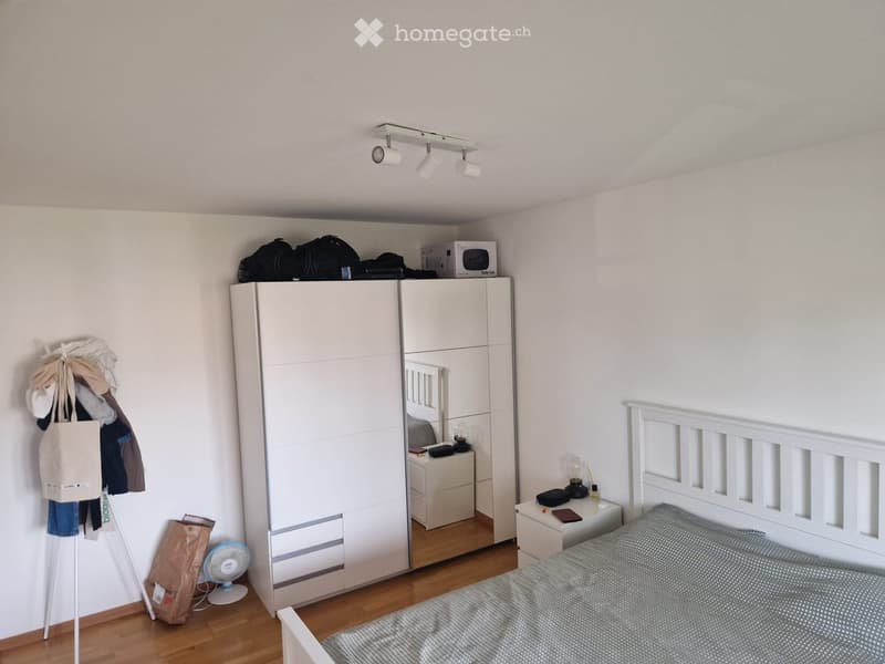 A modern bedroom with a white bed, wardrobe, nightstand, and a fan. The room features wooden flooring and white walls.