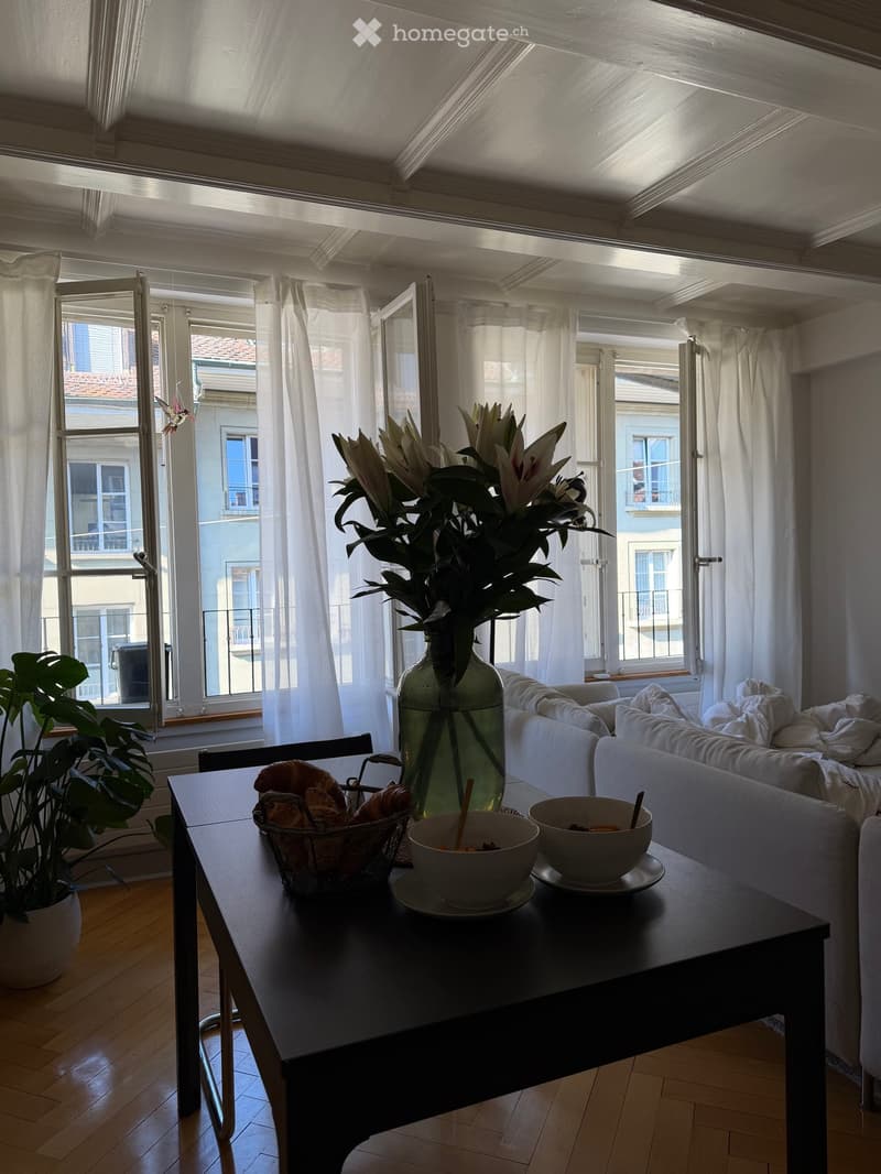 Bright living room with wooden floors, large windows, white curtains, a sofa, a coffee table with bowls and a vase of flowers, and a plant in a pot.