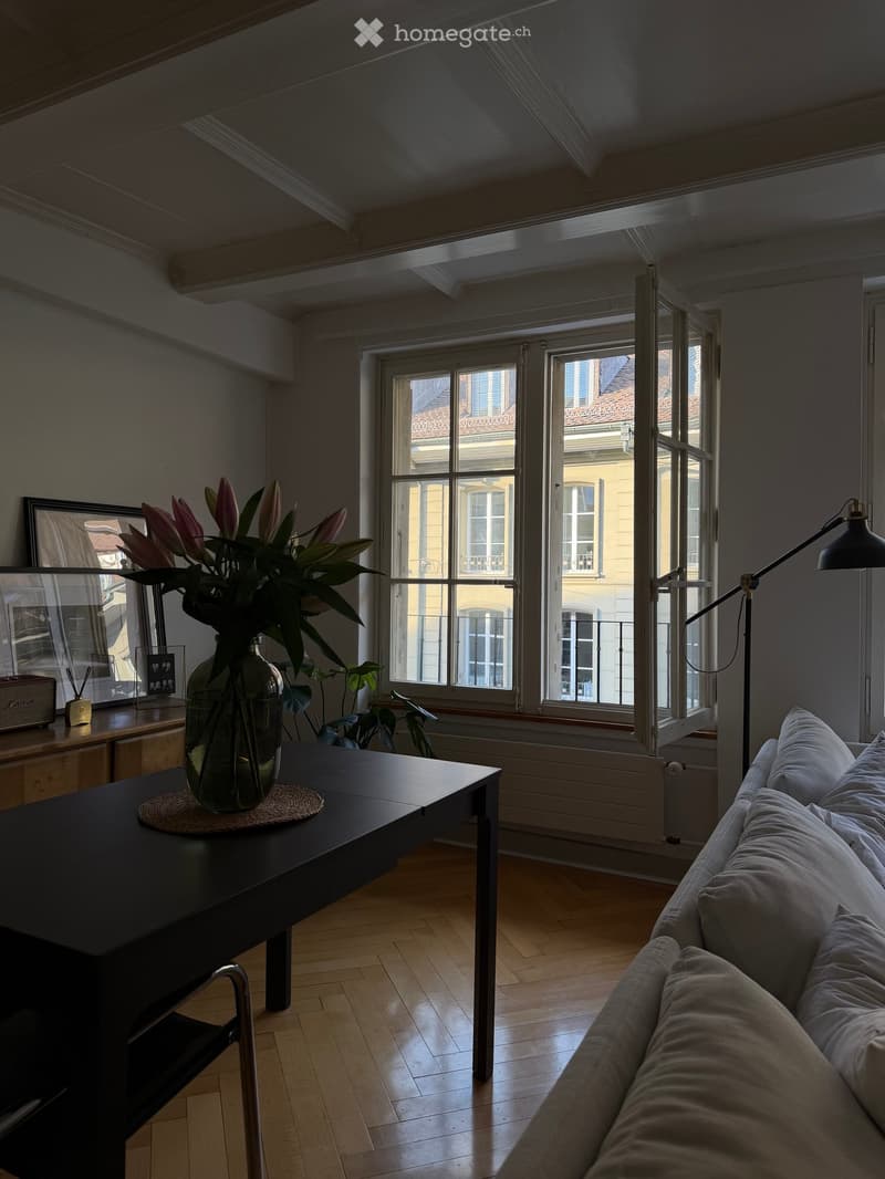 Living room with wooden floor, large windows, sofa, coffee table with vase of flowers, floor lamp, and picture frame on side table.