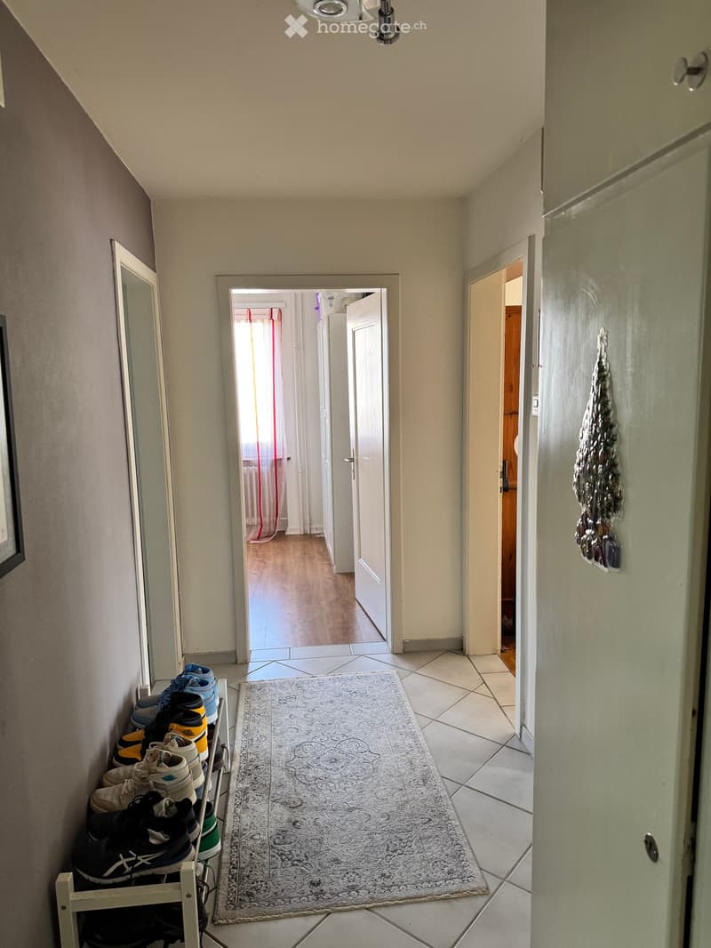 Hallway with tiled floor, a small rug, and a shoe rack holding multiple pairs of shoes. Open doors lead to other rooms.