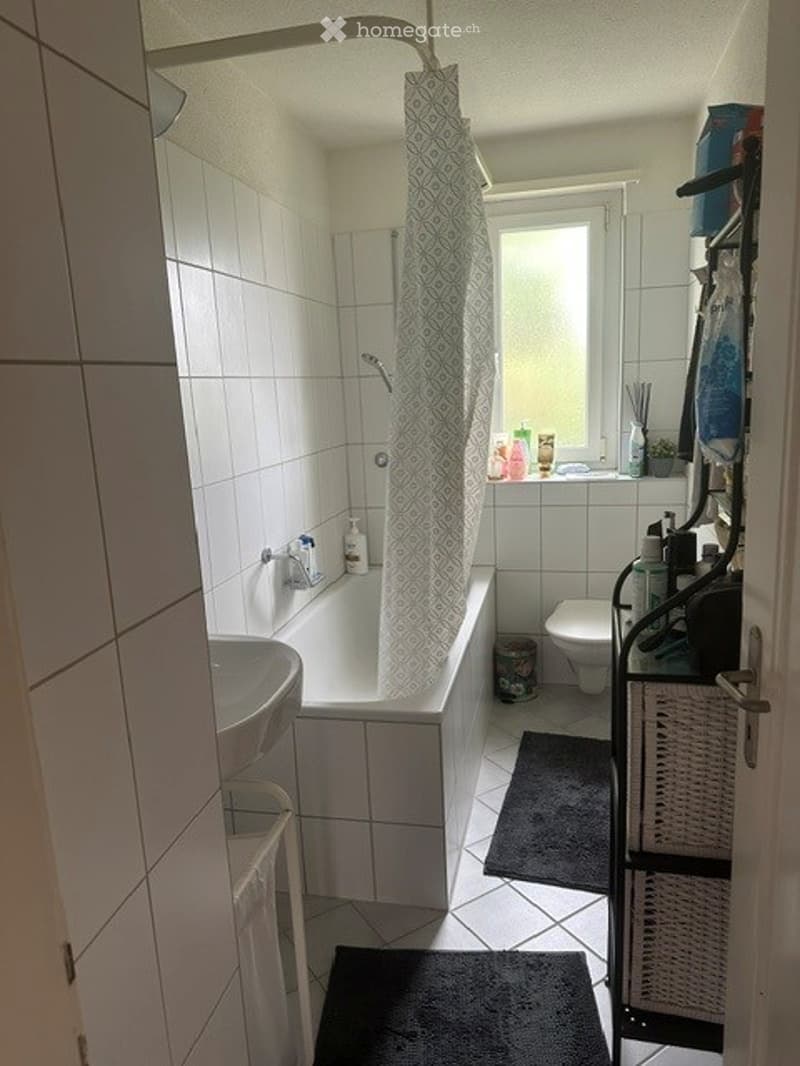 A modern bathroom with white tiles, a bathtub, toilet, sink, shower curtain, and a small shelf with toiletries.