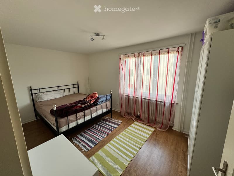 A small bedroom with a metal-framed bed, pillows, blankets, and two rugs on a wooden floor. The room has light-colored walls and a window with sheer curtains.