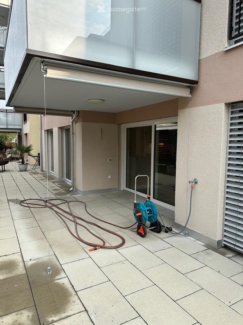 Outdoor patio area with tiled flooring, glass-enclosed balcony, hose reel, and potted plant visible in the background.