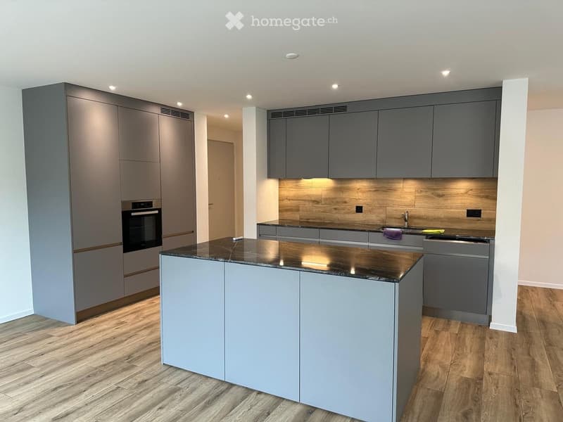 Modern kitchen with grey cabinets, a central island, and a dark countertop. Features an oven, sink, and faucet.
