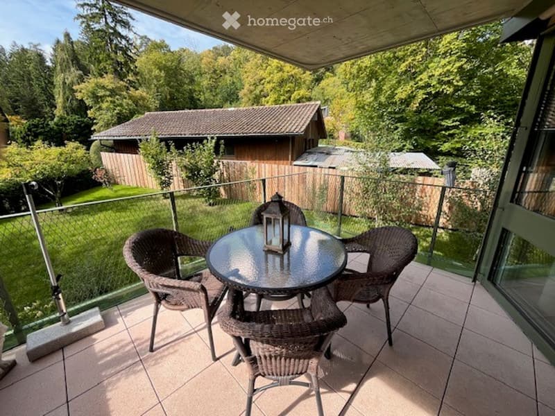 Balcony with a round glass table, four wicker chairs, and a lantern centerpiece, overlooking a green garden with trees and a wooden shed.