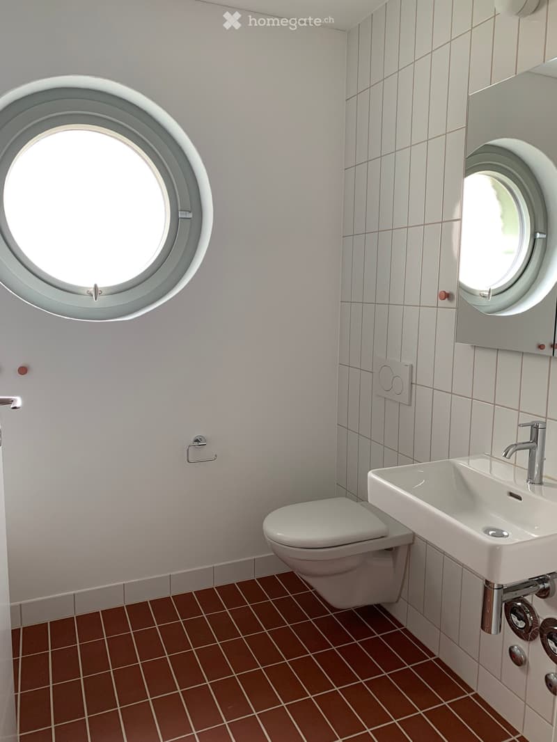 Modern bathroom with white tiled walls, brown tiled floor, a toilet, a sink with a modern faucet, and a mirror with a round window above it.