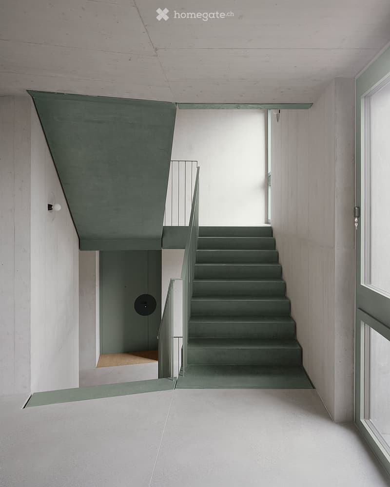 Modern hallway with a staircase leading upwards, featuring grey walls, a metal railing, and a concrete floor.