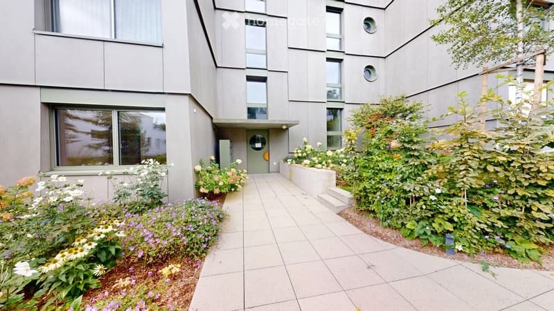 Modern building entrance with a tiled pathway, surrounded by well-maintained garden beds with colorful flowers and shrubs.