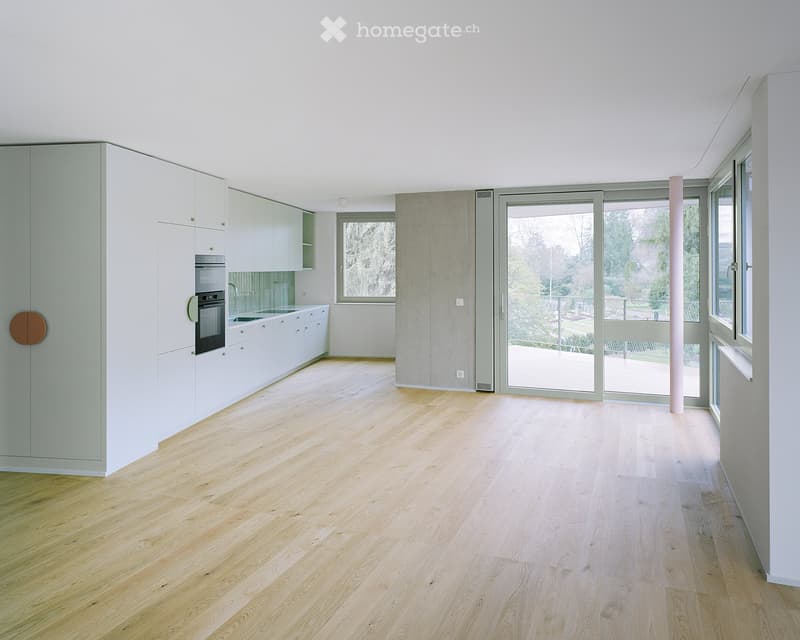 Open-plan space with light wooden flooring, white cabinetry, and large windows and sliding doors leading to an outdoor area.