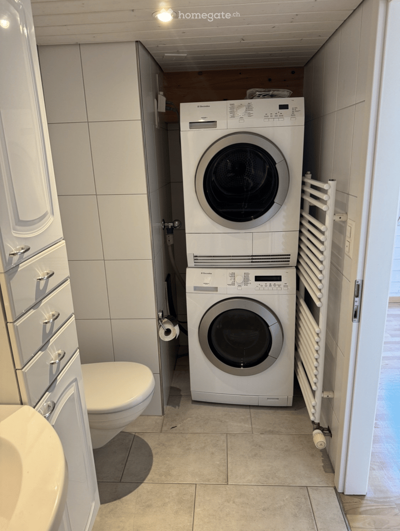 Modern bathroom with a toilet, washing machine, dryer, and radiator. The space is compact with white tiled walls and a tiled floor.