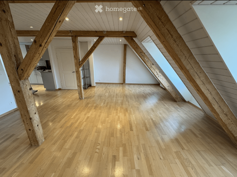 An empty attic space with wooden beams and exposed wooden floorboards, featuring large skylight windows and white walls.