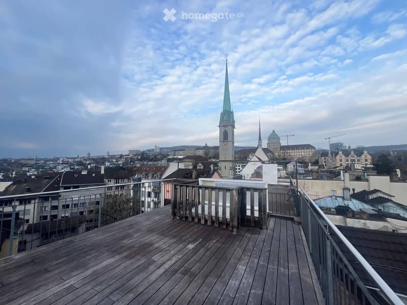 Wooden balcony with metal railing, overlooking a cityscape with historic buildings and a church spire.