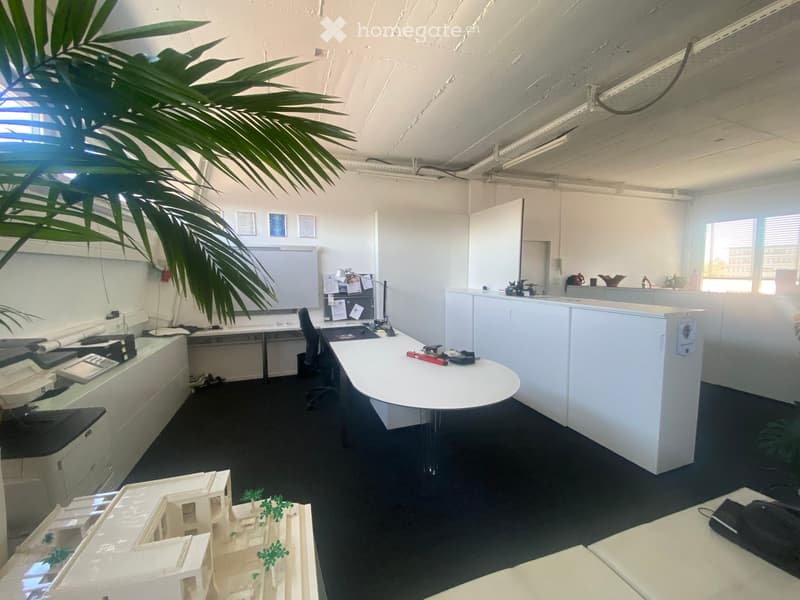 Office space with desks, chairs, a large screen, printer, model, and a plant. The room is well-lit with white walls and a dark carpeted floor.