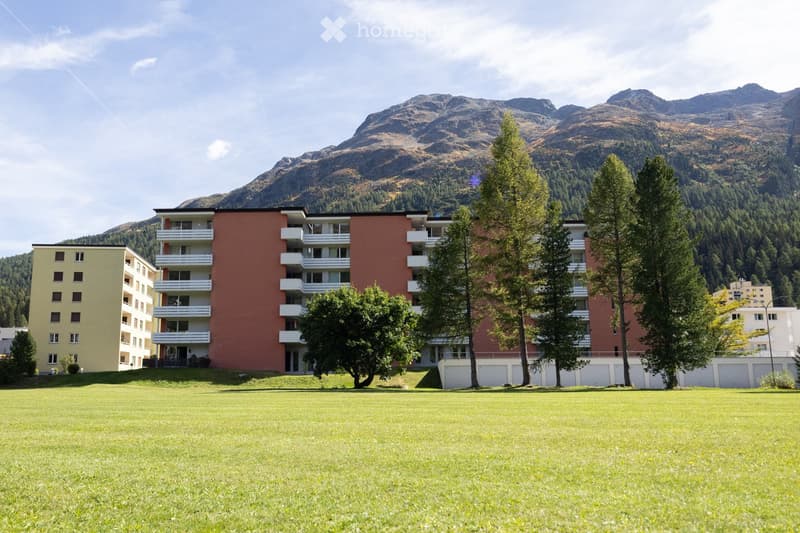 Multi-story apartment building with balconies, surrounded by trees and a grassy lawn, with mountain backdrop.