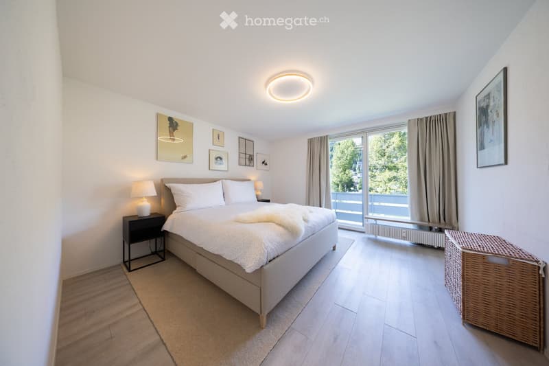 Modern bedroom with a large bed, nightstand, lamp, and artwork on the walls. The room features wooden flooring, large windows with curtains, and a wicker basket near the window.