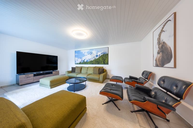 Modern living room with a green sectional sofa, coffee table, ottomans, leather chairs, and a large television. The room features white walls, a white shag rug, and several framed artworks.