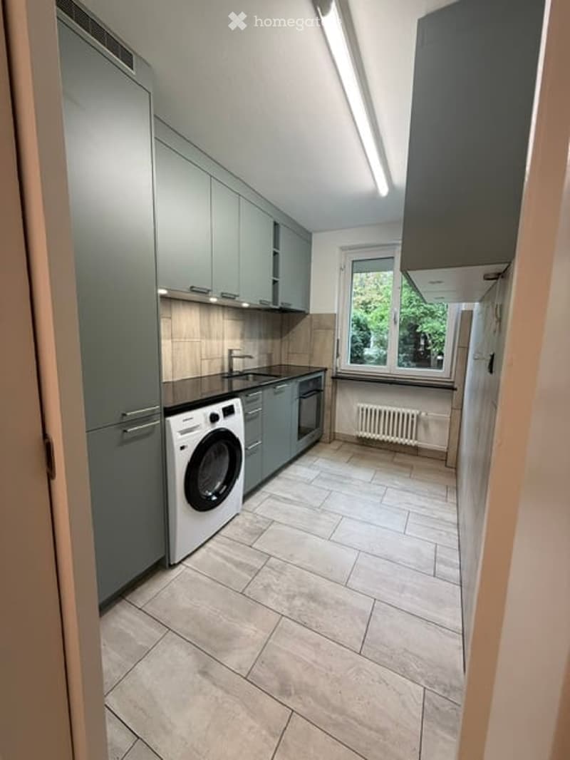 Modern kitchen with grey cabinets, a white washing machine, a sink with a faucet, and an oven. The floor is tiled, and there is a window with a view of greenery.