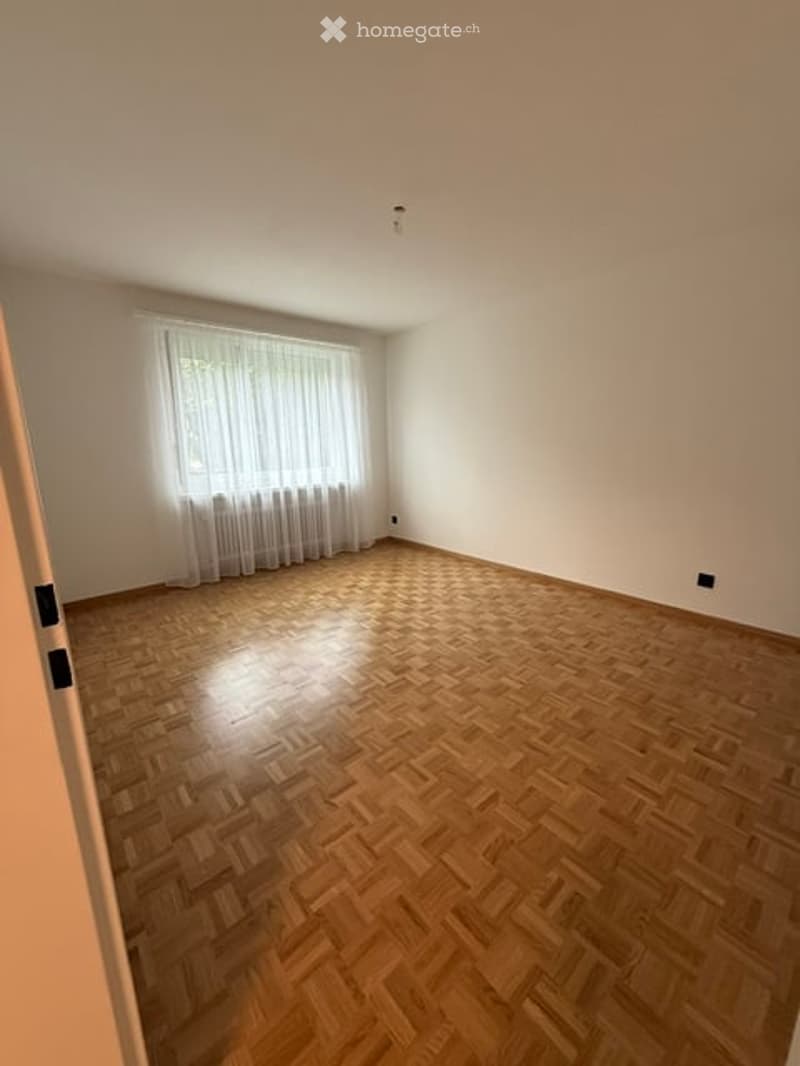 Empty room with wooden parquet flooring, white walls, and a large window with white curtains.