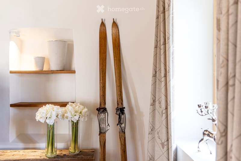 A cozy, well-lit room with wooden skis leaning against the wall, two vases with white flowers on a wooden shelf, and a decorative candle holder on the right side.