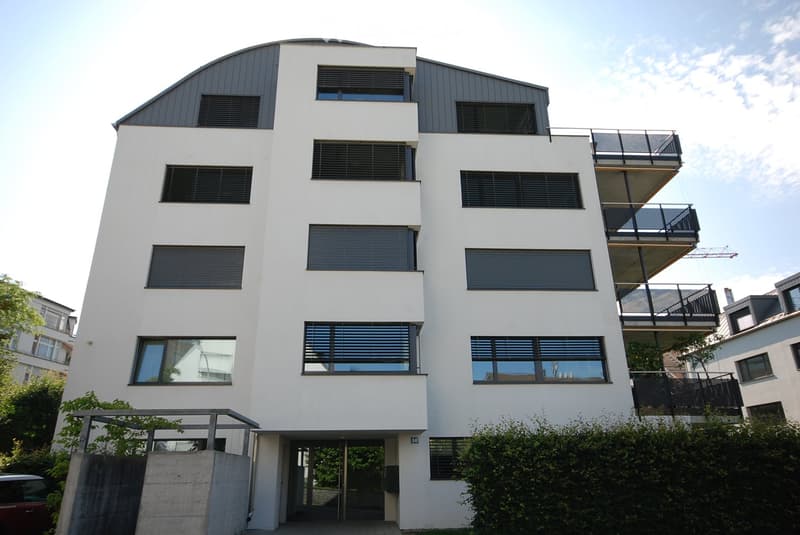 Modern, multi-story building with white facade, large windows, and balconies. The structure features a sleek, contemporary design with a combination of flat and sloped roofing.
