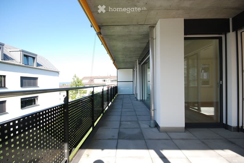 Modern balcony with tiled floor, black metal railing, glass doors leading to interior, and a view of neighboring buildings.