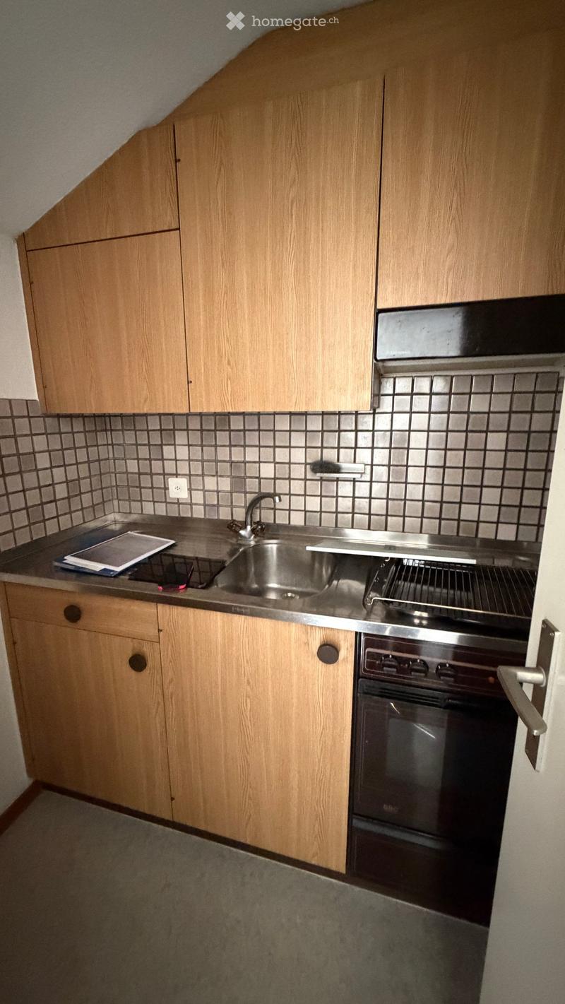 Compact kitchen with wooden cabinets, stainless steel sink, and oven. Countertop has a few items including a tray and papers.