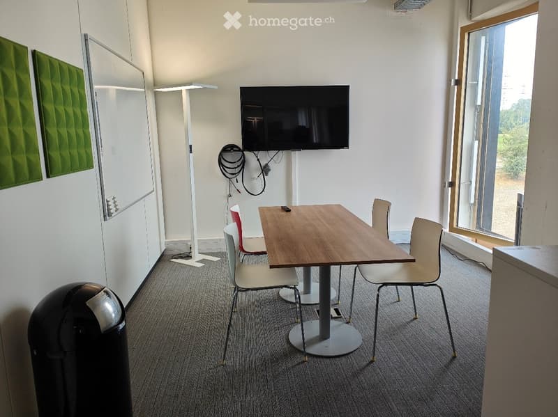 A modern, well-lit room with a wooden table, several chairs, a mounted screen, a floor lamp, and a trash bin. The room features large windows and acoustic panels on the walls.