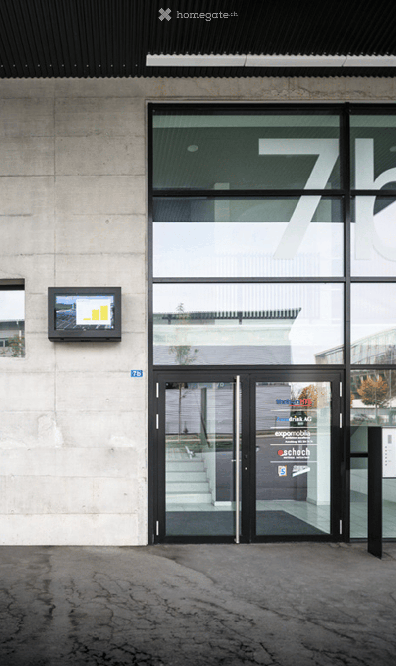 Modern building entrance with large glass doors, a digital display screen mounted on the wall, and a concrete facade.